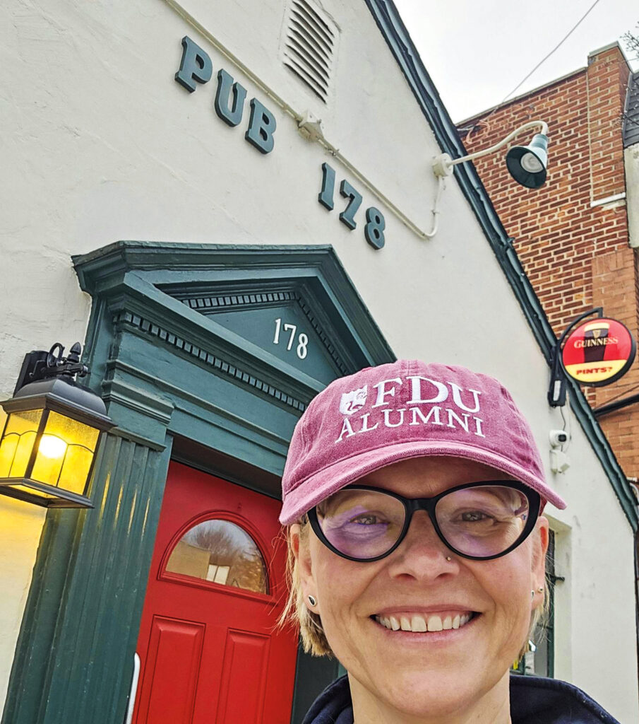 A woman stands with a building behind her wearing a baseball cap and glasses.