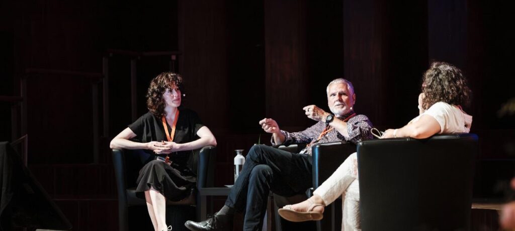 Three people participate in a panel discussion on a stage, seated in black armchairs. On the left, a woman in black with an orange lanyard listens, while a man in the center gestures as he speaks to a woman in white on the right. A stage monitor sits on the floor in the foreground.