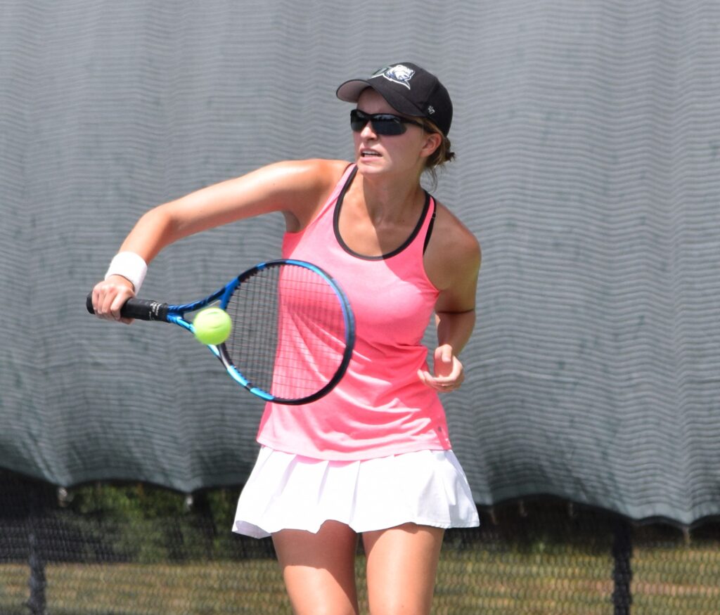 A woman swinging a tennis racket at a tennis ball wearing a pink and white tennis dress, sunglasses and a baseball cap.