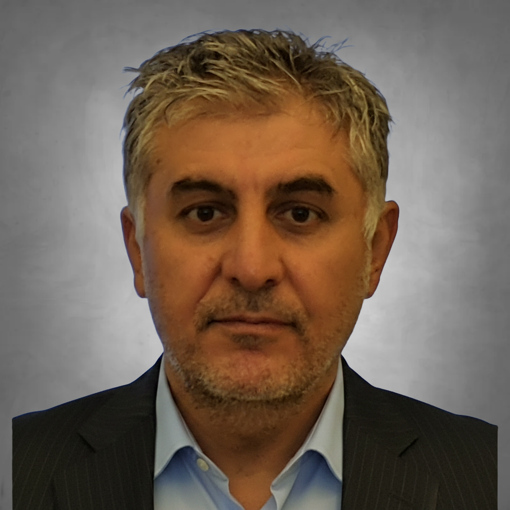 A professional headshot of a man with short salt-and-pepper hair and light facial stubble. He is wearing a dark suit jacket over a light blue collared shirt and is photographed against a neutral gray background with a serious, professional expression.