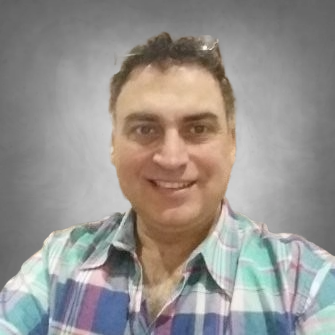 A professional headshot of a person with dark hair wearing a plaid button-up shirt in shades of blue, green, and white. They are smiling at the camera and photographed against a neutral gray background.