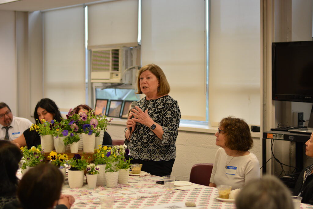 A woman in a black and white patterned top speaks into a microphone at a meeting, standing behind a table filled with small potted flowers while seated attendees listen.