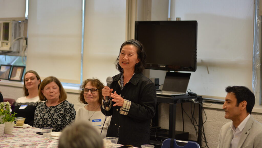 A woman in a black jacket stands and speaks into a microphone at a meeting, while other attendees sit around a table listening. A TV monitor is visible in the background.