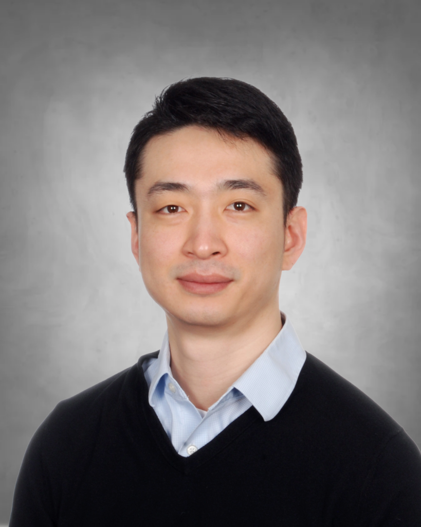 A professional headshot of a person with short dark hair. They are wearing a black v-neck sweater over a light blue collared dress shirt. They have a gentle, pleasant smile and are photographed against a neutral gray background.