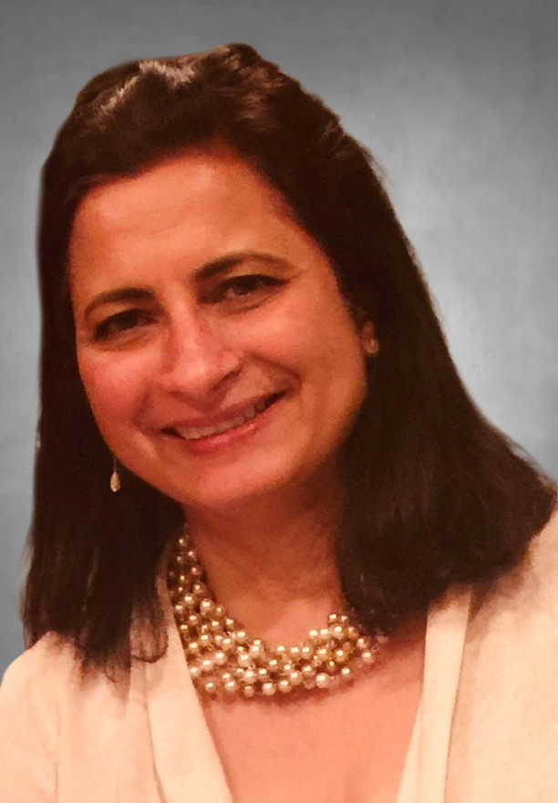 A professional headshot of a woman with dark brown shoulder-length hair. She is wearing a white or cream-colored blouse and a multi-strand pearl necklace. She has a warm smile and is photographed against a light blue-gray background.