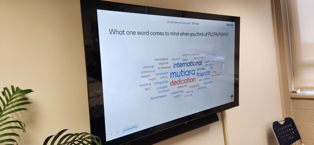 A wall-mounted TV displays an upside-down Mentimeter word cloud. The prompt asks, "What one word comes to mind when you think of PLCPA/PEAPS?" with prominent words including "international", "mutiara", and "dedication".