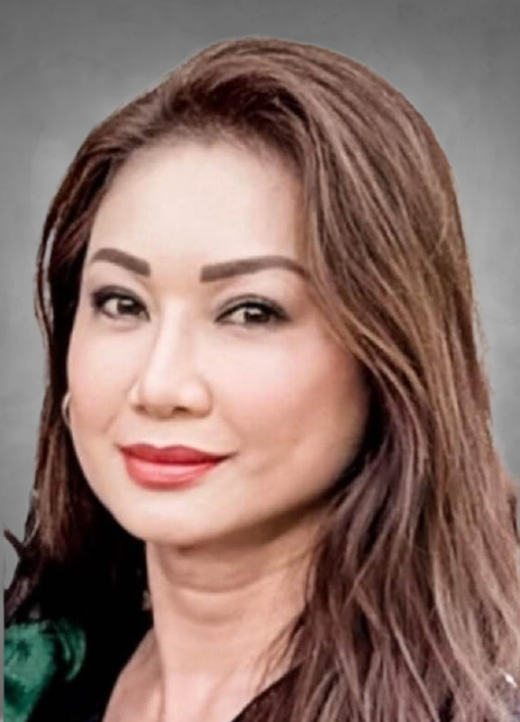 A professional headshot of a woman with long brown hair with lighter highlights. She is wearing professional attire and has a warm, pleasant smile. She is photographed against a neutral gray background.