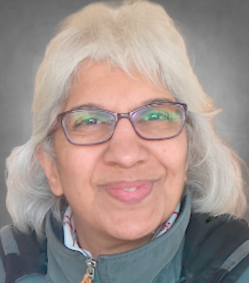 A professional headshot of a person with shoulder-length white/gray hair wearing glasses with dark frames. They are dressed in a teal zip-up jacket and smiling at the camera against a neutral gray background.