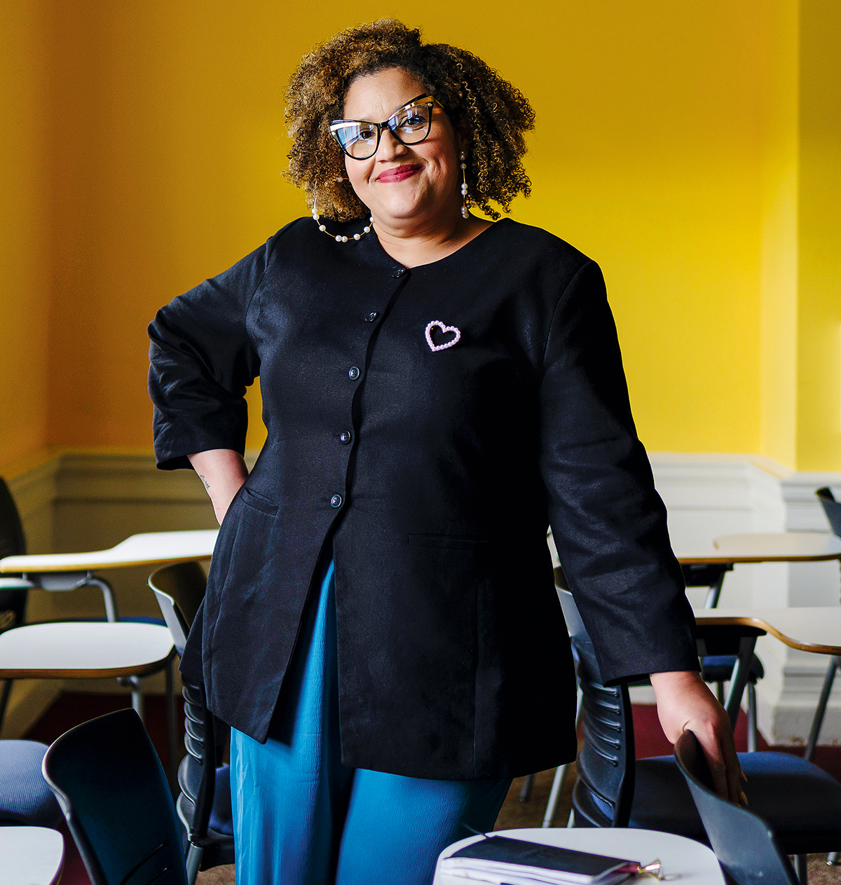 A woman wearing glasses, smiles and puts her hands on her hip in a classroom.