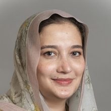 A professional headshot of a woman wearing a light-colored patterned headscarf. She has a gentle smile and is photographed against a neutral gray background.