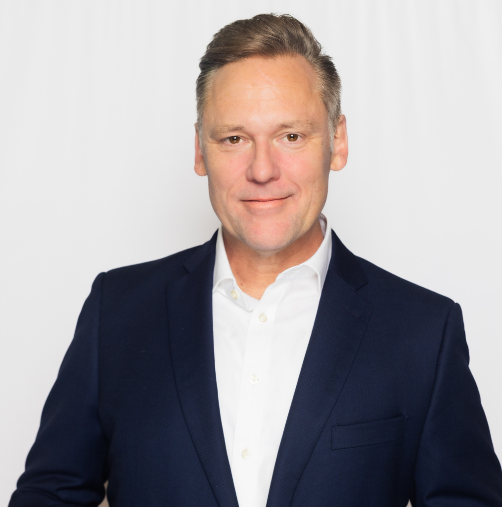 Professional headshot of a man smiling slightly. He is wearing a white button-down shirt and a navy blue blazer against a plain white background.