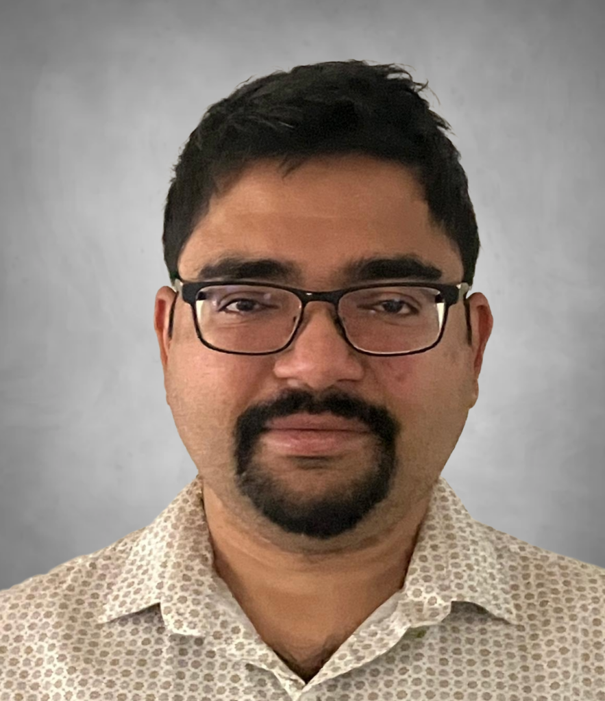 A professional headshot of a man with short dark hair, wearing black-framed glasses and a goatee. He is wearing a light-colored button-up shirt with a small geometric or dotted pattern and is photographed against a neutral gray background with a friendly expression.