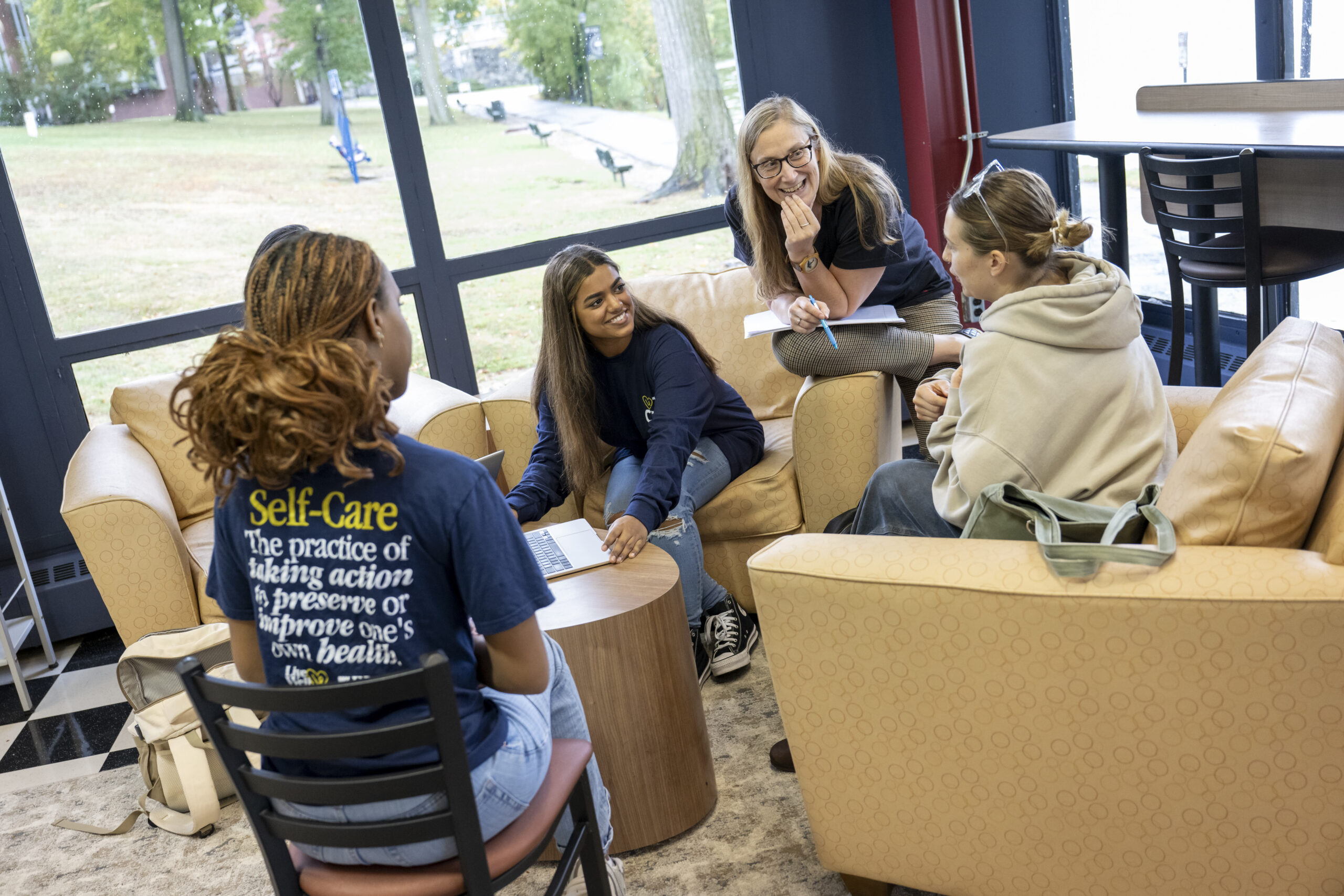 Four people are seated in a circle in a comfortable lounge area with large windows overlooking a grassy campus. A woman with glasses, holding a notebook and pen, smiles as she leans forward, engaging with three other women. One person sits with a laptop on a small round table, while another, seen from behind, wears a blue t-shirt with the text 
