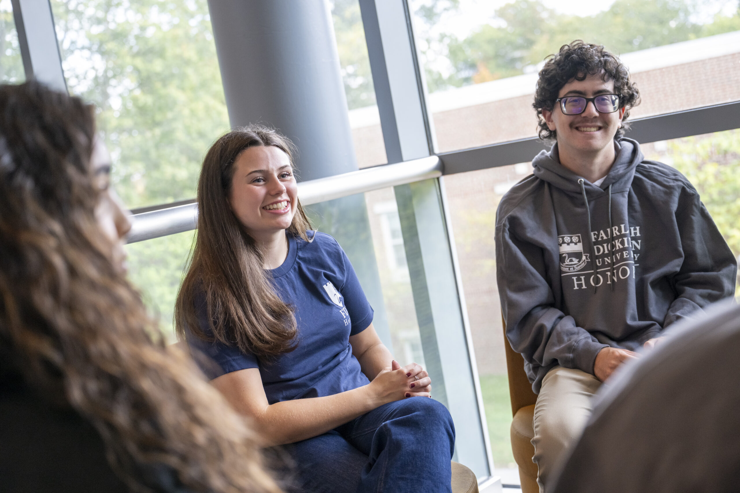 A group of students sits in a circle near a large window, engaged in conversation. A man with curly hair and glasses wears a grey hoodie with the text 