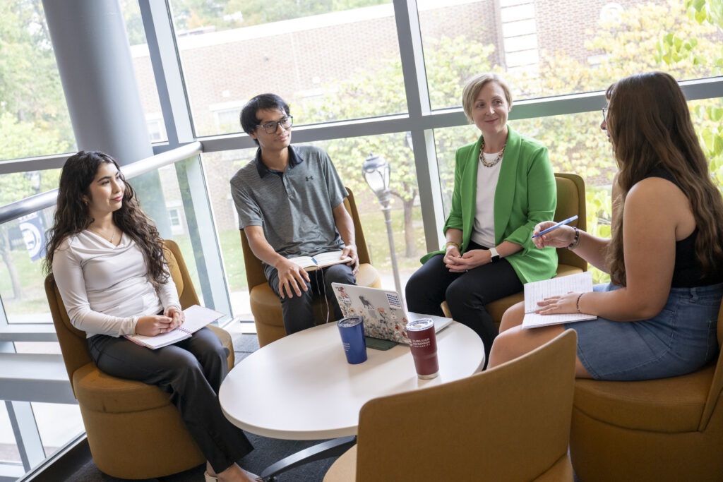 Four people sit in a semi-circle on mustard-yellow chairs around a small white table in a bright room with large windows. They appear engaged in a discussion, holding notebooks while a laptop and tumblers rest on the table.