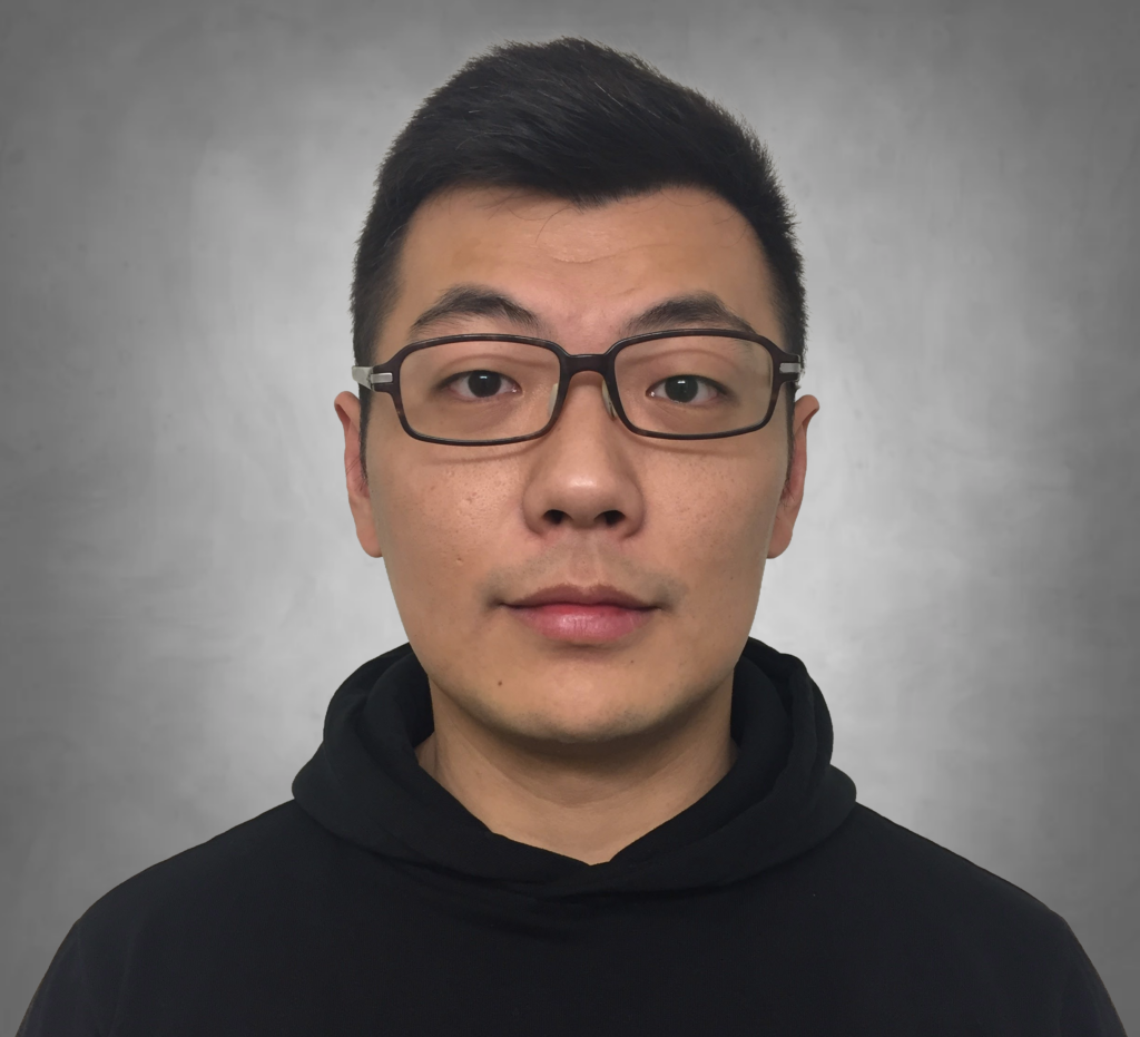 A professional headshot of a person with short dark hair wearing dark-framed glasses. They are wearing a black hoodie or high-necked garment and are photographed against a neutral gray background with a neutral expression.