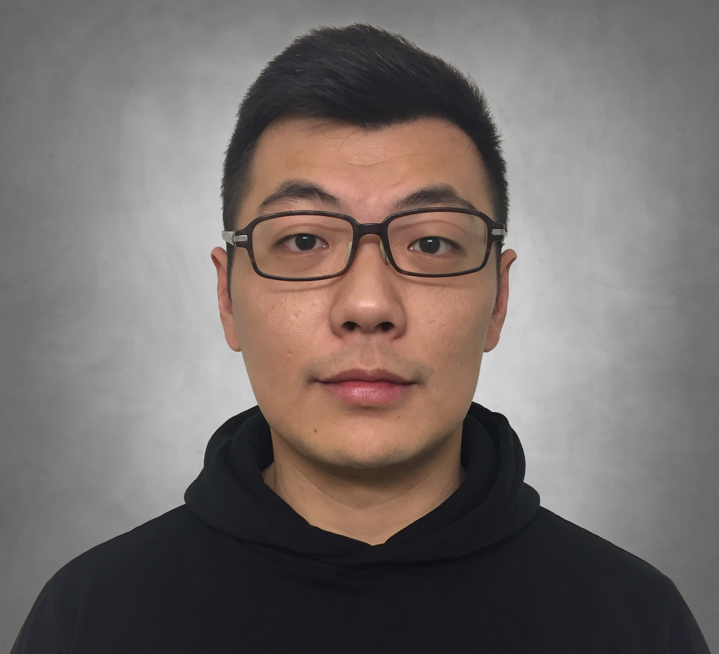 A professional headshot of a person with short dark hair wearing dark-framed glasses. They are wearing a black hoodie or high-necked garment and are photographed against a neutral gray background with a neutral expression.