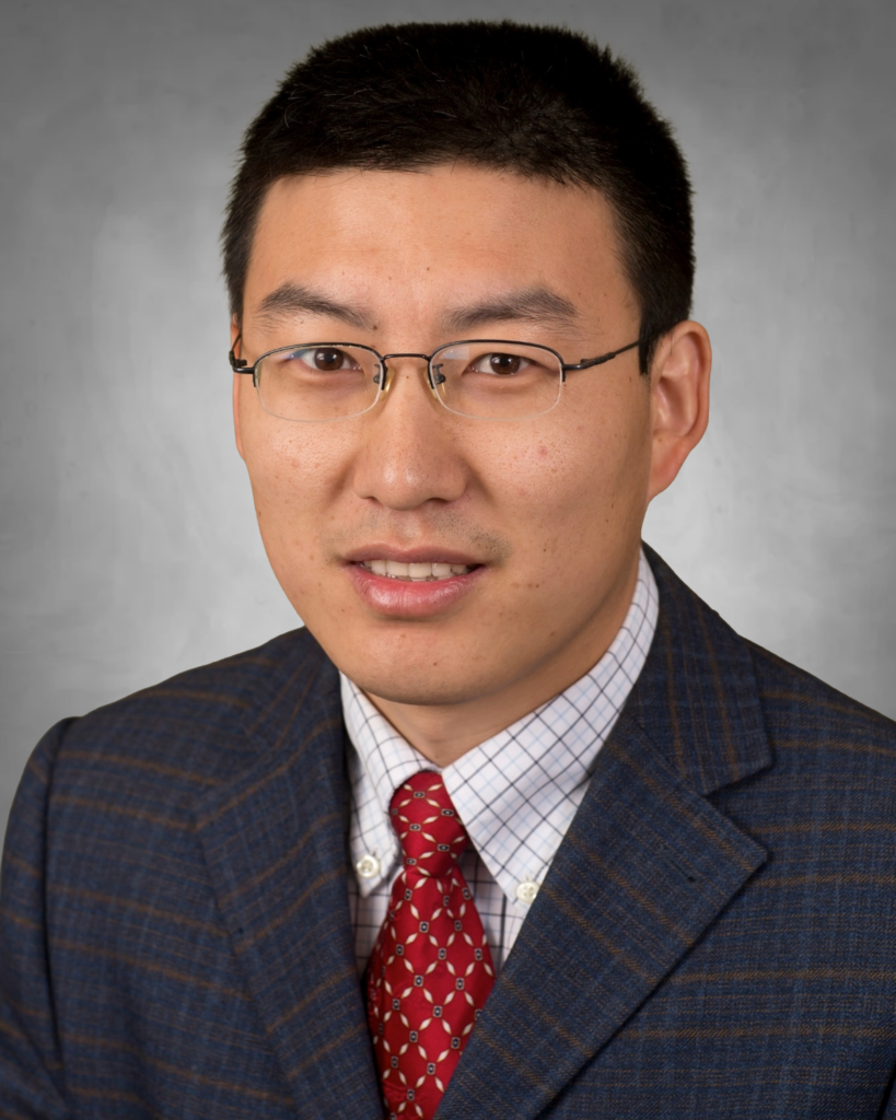 A professional headshot of a man with short black hair wearing glasses. He is dressed in a blue plaid blazer, a light checkered dress shirt, and a red patterned tie. He has a slight smile and is photographed against a neutral gray background.