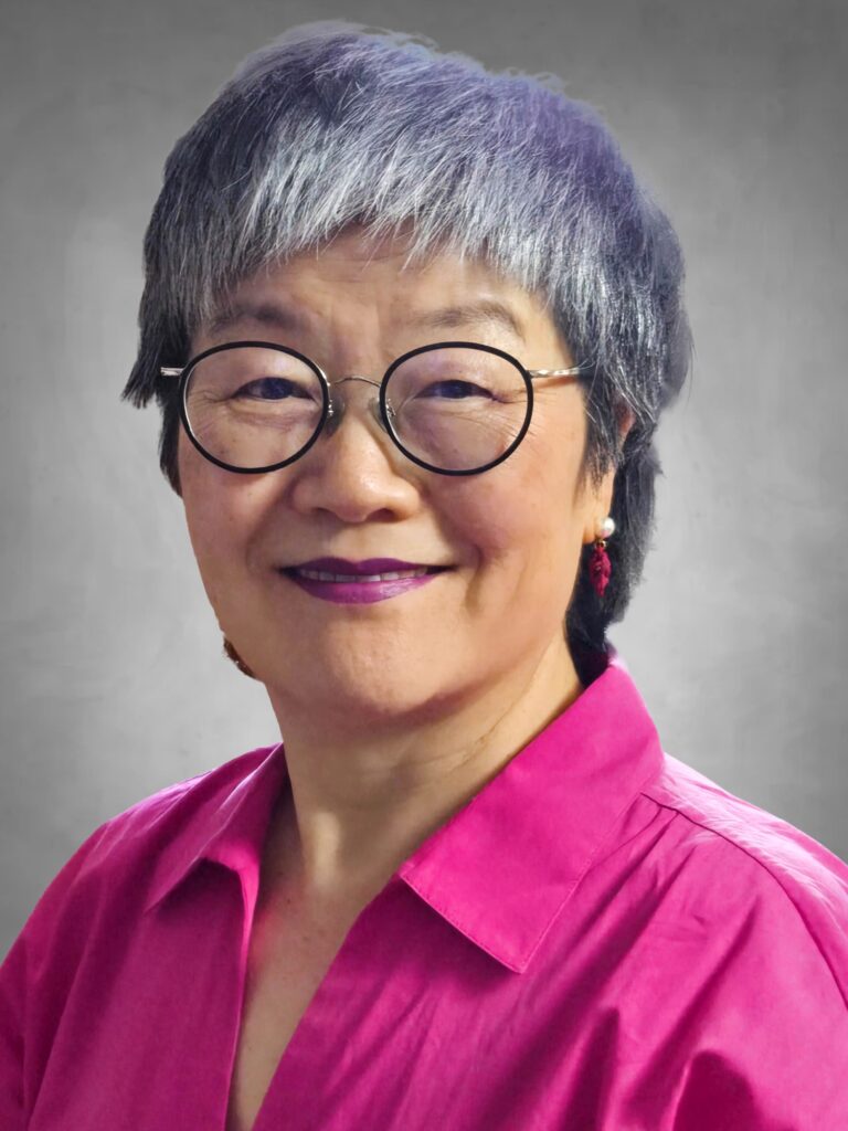 A professional headshot of a person with short gray hair wearing round black-framed glasses. They are dressed in a bright magenta collared shirt and have purple lipstick. They are smiling warmly at the camera against a neutral gray background.