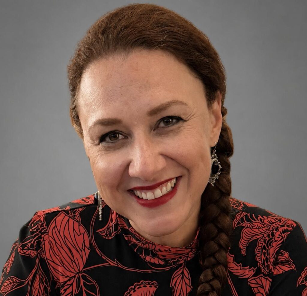 A professional headshot of a woman with brown hair styled in a side braid. She is wearing a black blouse with a red and orange floral pattern and decorative drop earrings. She is smiling warmly at the camera against a neutral gray background.