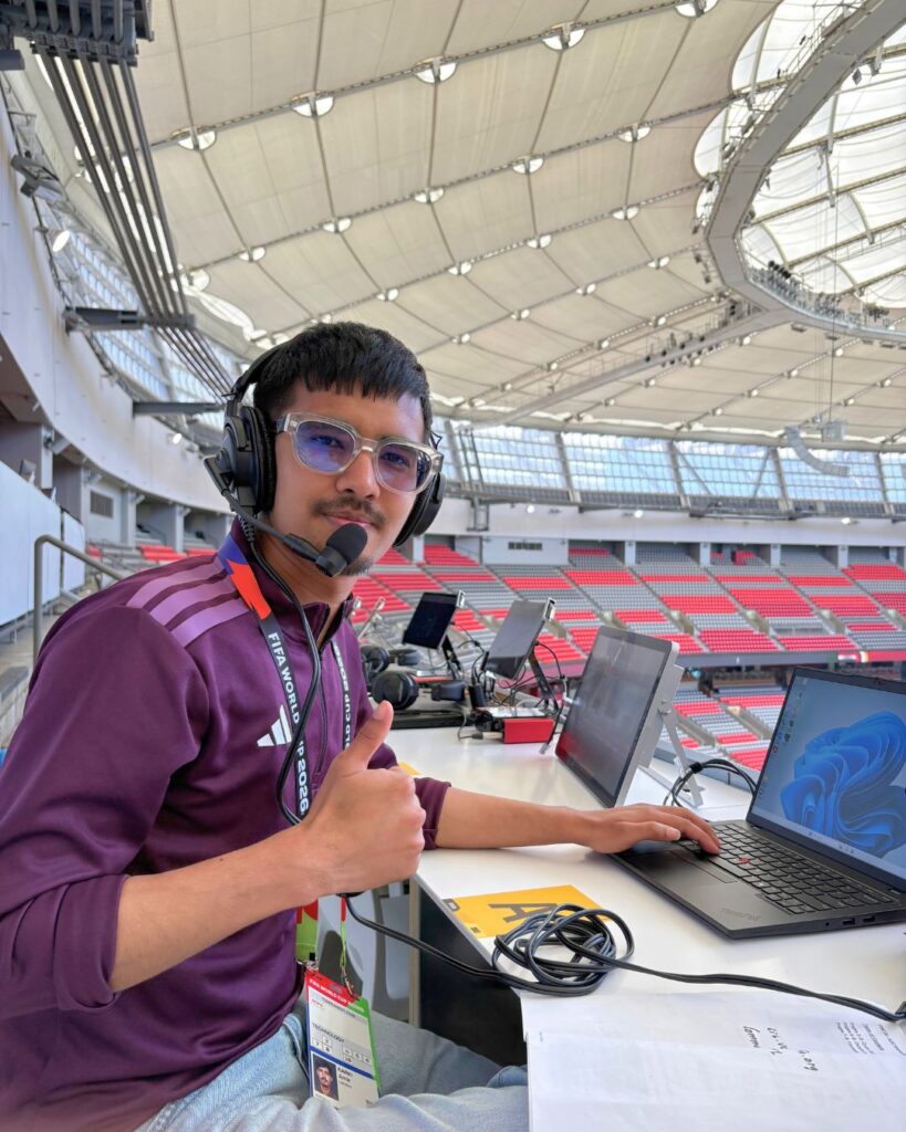a young man wears a headset and gives a thumbs-up to the camera. he works on a laptop inside of a stadium.
