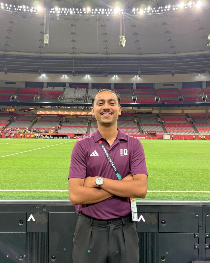 a young man smiles at the camera, with his arms crossed. he wears a red polo and stands inside of a stadium.