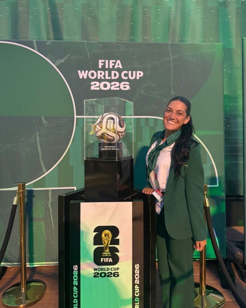 a woman wears a green suit and a lanyard. she stands next to a soccer ball labelled "official match ball." the sign above her reads "FIFA World Cup 2026"