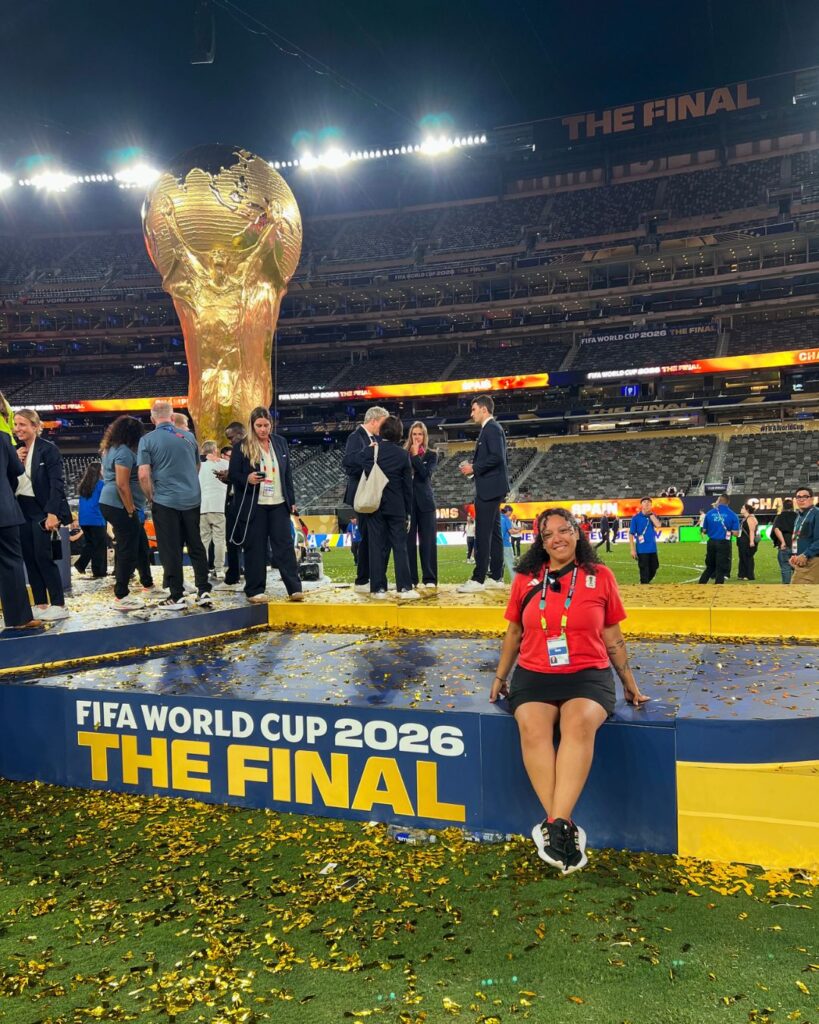 a young woman sits on the FIFA World Cup 2026 The Final stage. she wears a red shirt. there is a statue of the World Cup behind her.