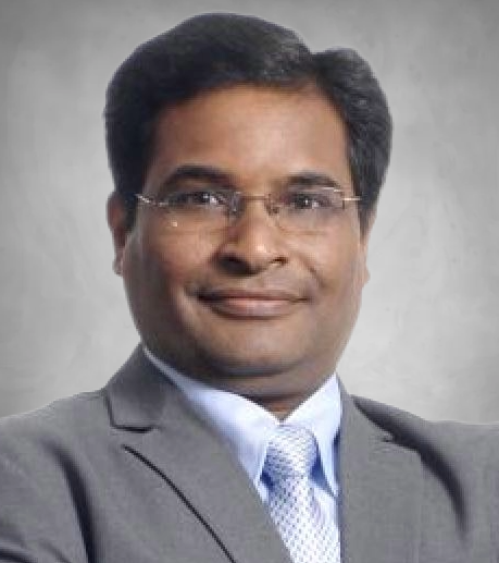A professional headshot of a person wearing glasses and business attire. They are dressed in a gray suit jacket, light blue dress shirt, and light colored tie. They have dark hair and a pleasant smile, photographed against a neutral gray background.