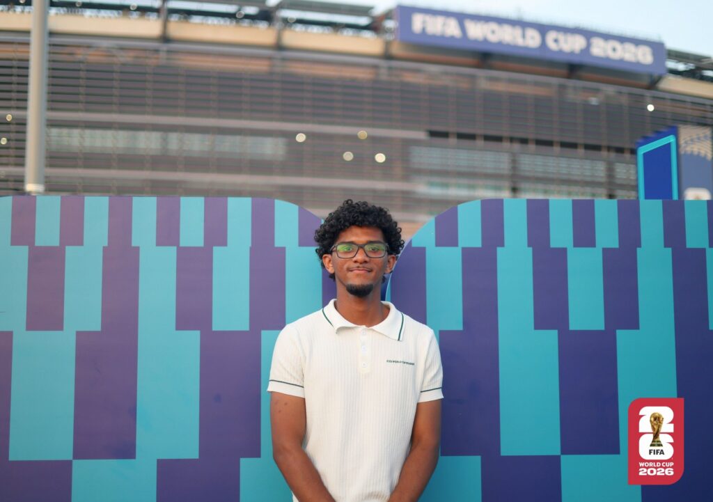 a college student smiles at the camera. he is in front of New York New Jersey Stadium.
