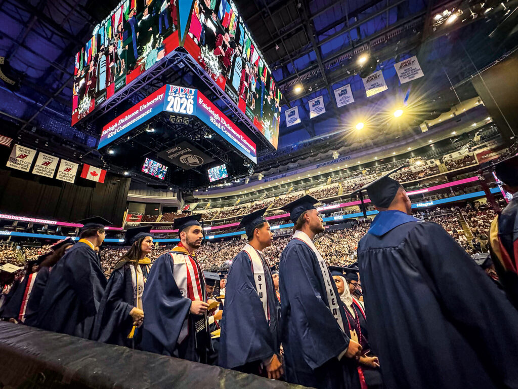 Students stand in a line waiting to walk across the stage the 2026 Commencement ceremony wearing navy blue caps and gowns.