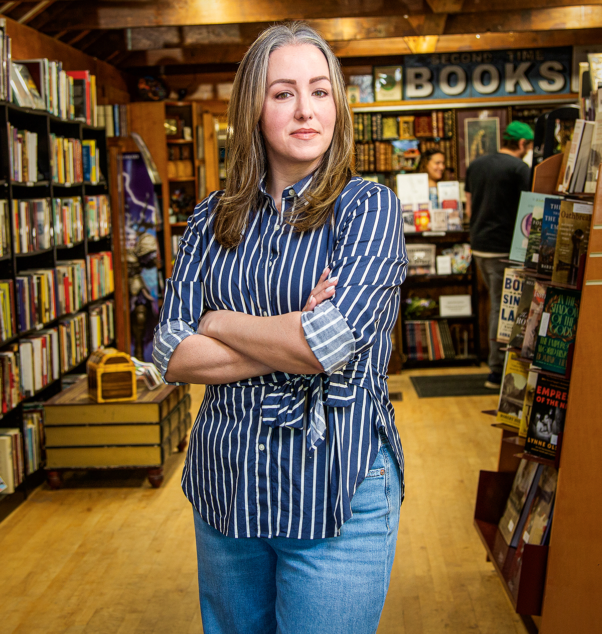 A woman crosses her arms and stands in a bookstore.