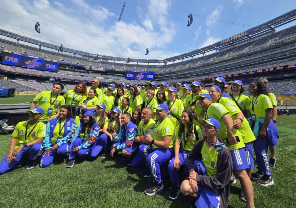 a group of volunteers poses for a photo. they stand on a soccer field. signs around them read "FIFA The Final."