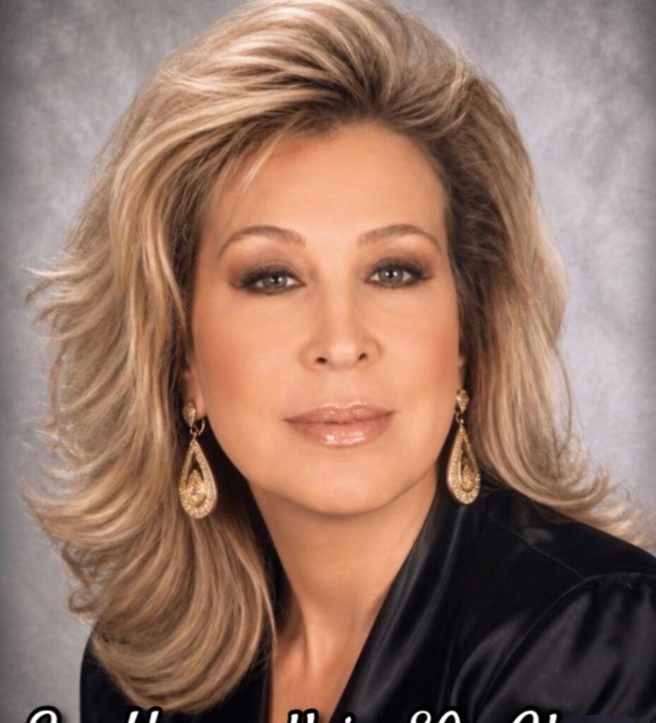 A woman with shoulder-length blonde hair, wearing a black top and gold earrings against a gray background.