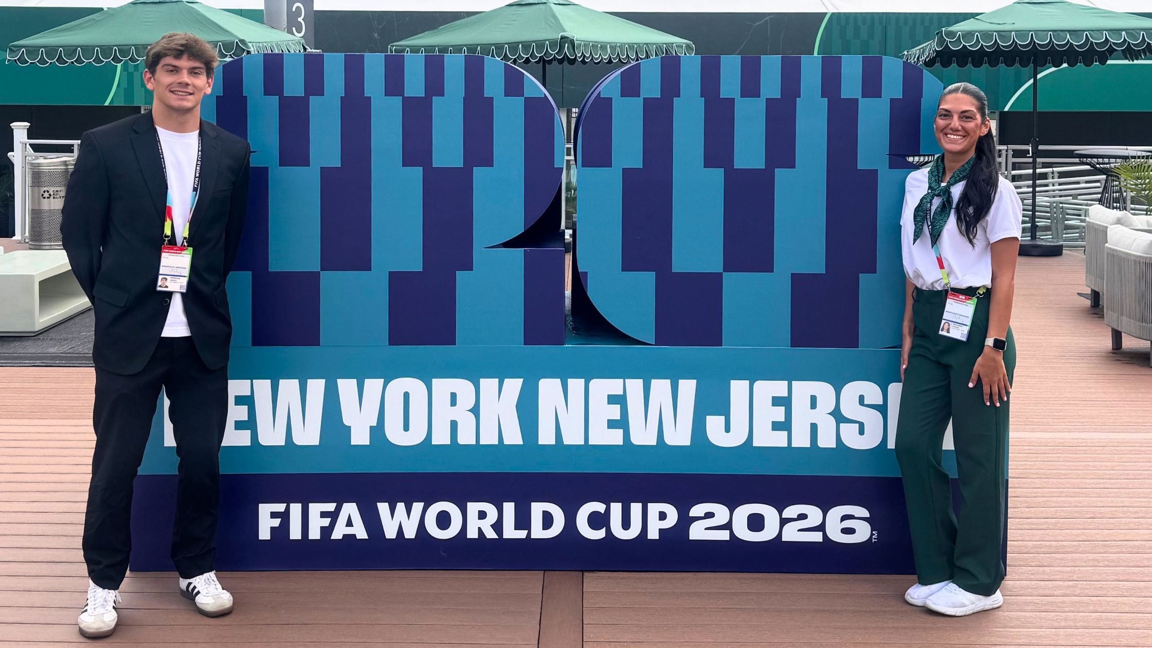 two FDU college students pose with a sign for the FIFA World Cup 2026. they are dressed in professional attire.