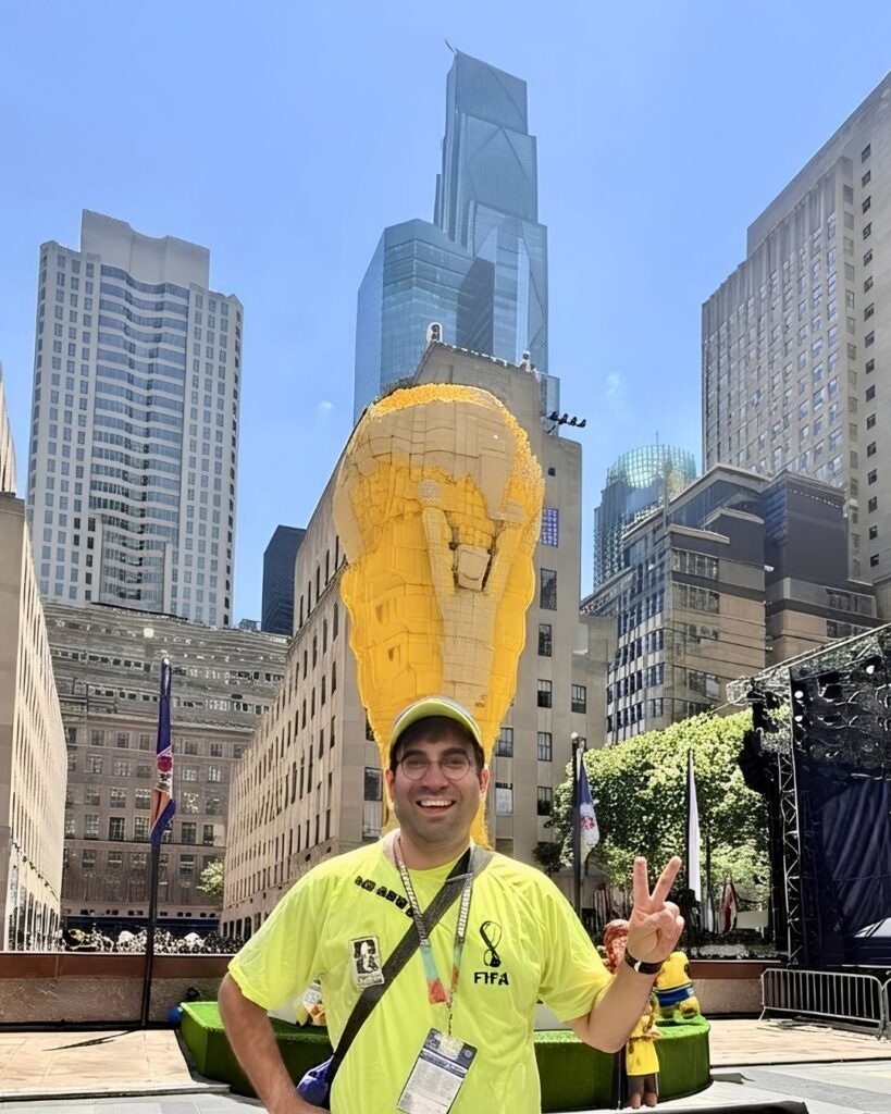 a man holds up a peace sign with his fingers and smiles at the camera. behind him there is a massive FIFA World Cup trophy made out of Legos.