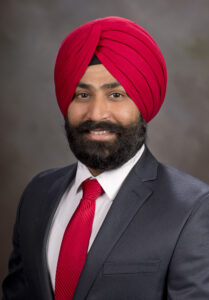 A professional headshot of a person wearing a red turban and business attire. They are dressed in a dark suit jacket, white collared shirt, and red striped tie that coordinates with the turban. The person has a full beard and is smiling at the camera against a neutral gray background.