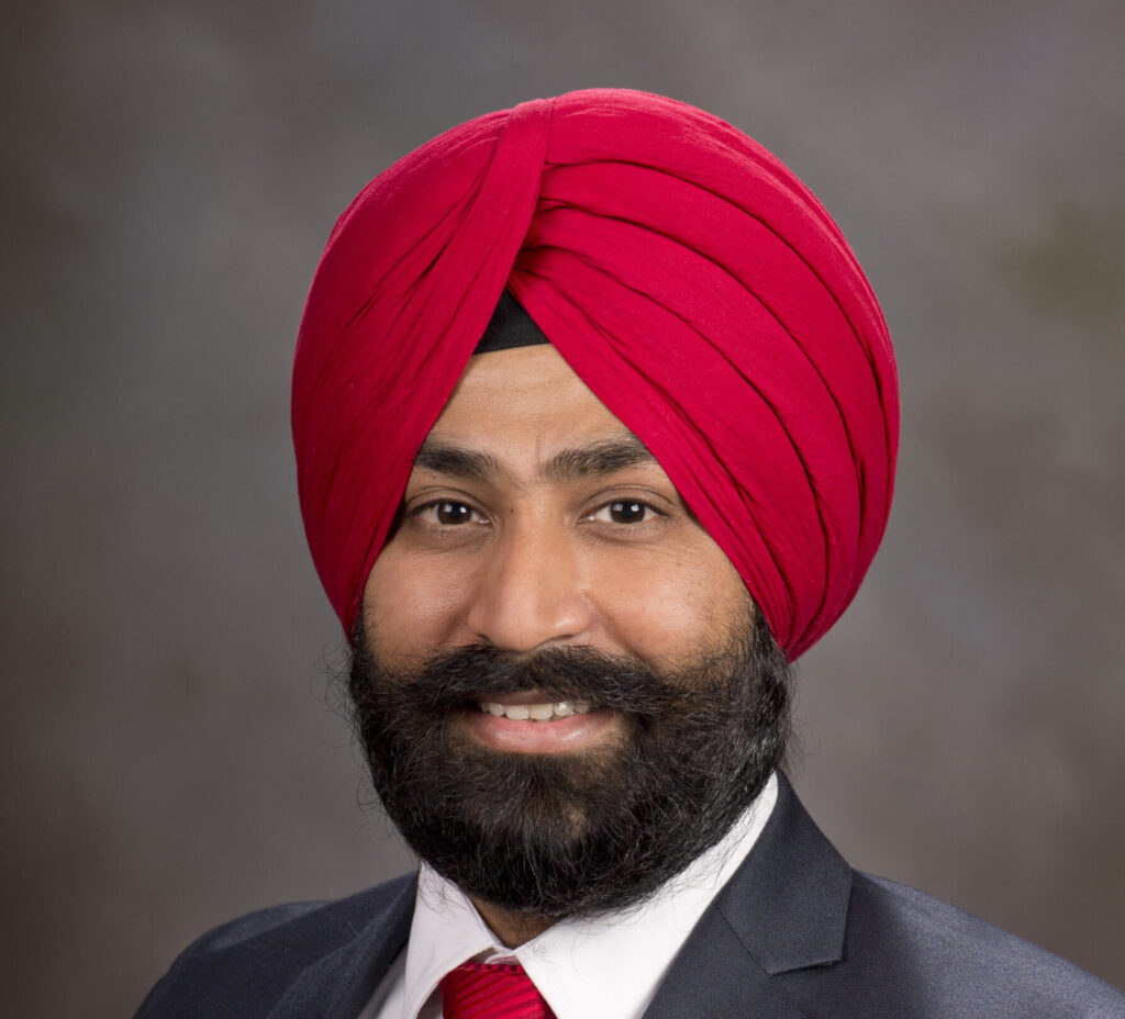 A professional headshot of a person wearing a red turban and business attire. They are dressed in a dark suit jacket, white collared shirt, and red striped tie that coordinates with the turban. The person has a full beard and is smiling at the camera against a neutral gray background.