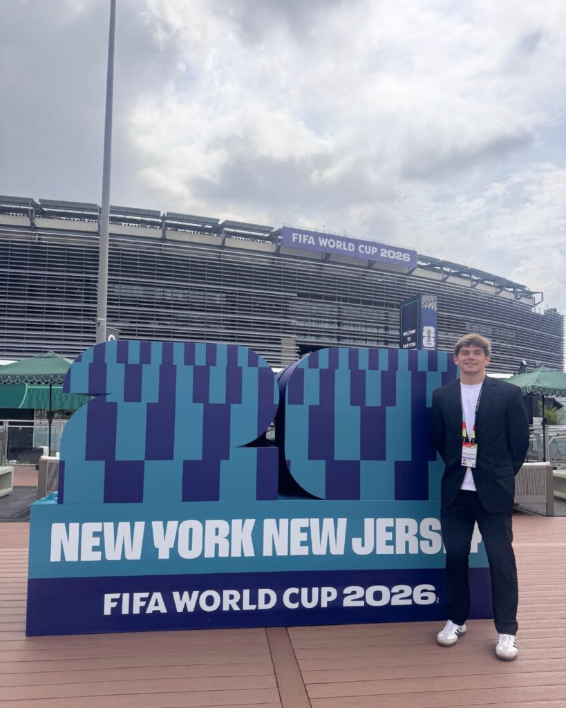 a young man poses in front of a FIFA World Cup 2026 sign. he wears a suit. New York New Jersey Stadium is behind him.