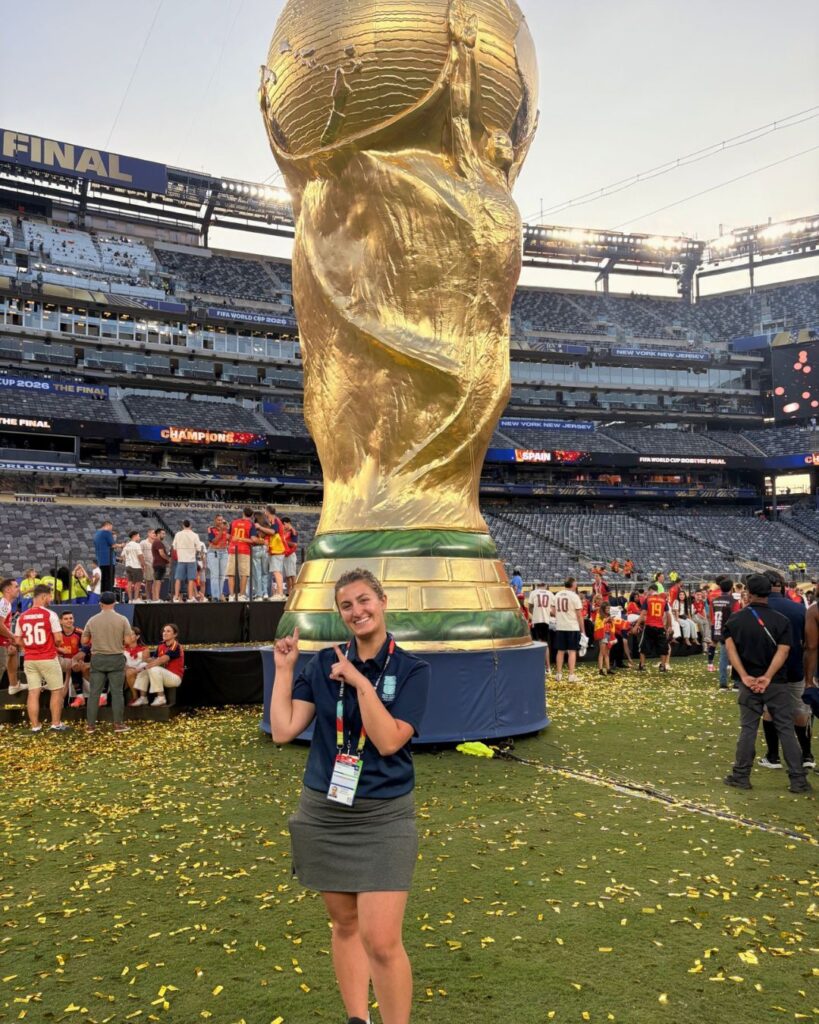 a young woman points to the statue of the World Cup behind her. she wears a polo and skirt uniform.