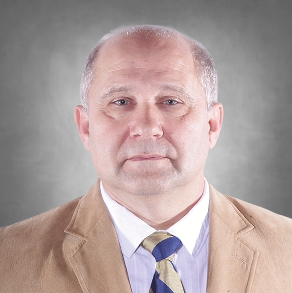 A professional headshot of a man with short hair. He is wearing a tan blazer, a light purple collared shirt, and a blue and gold striped tie. He looks directly at the camera with a neutral expression against a neutral gray background.