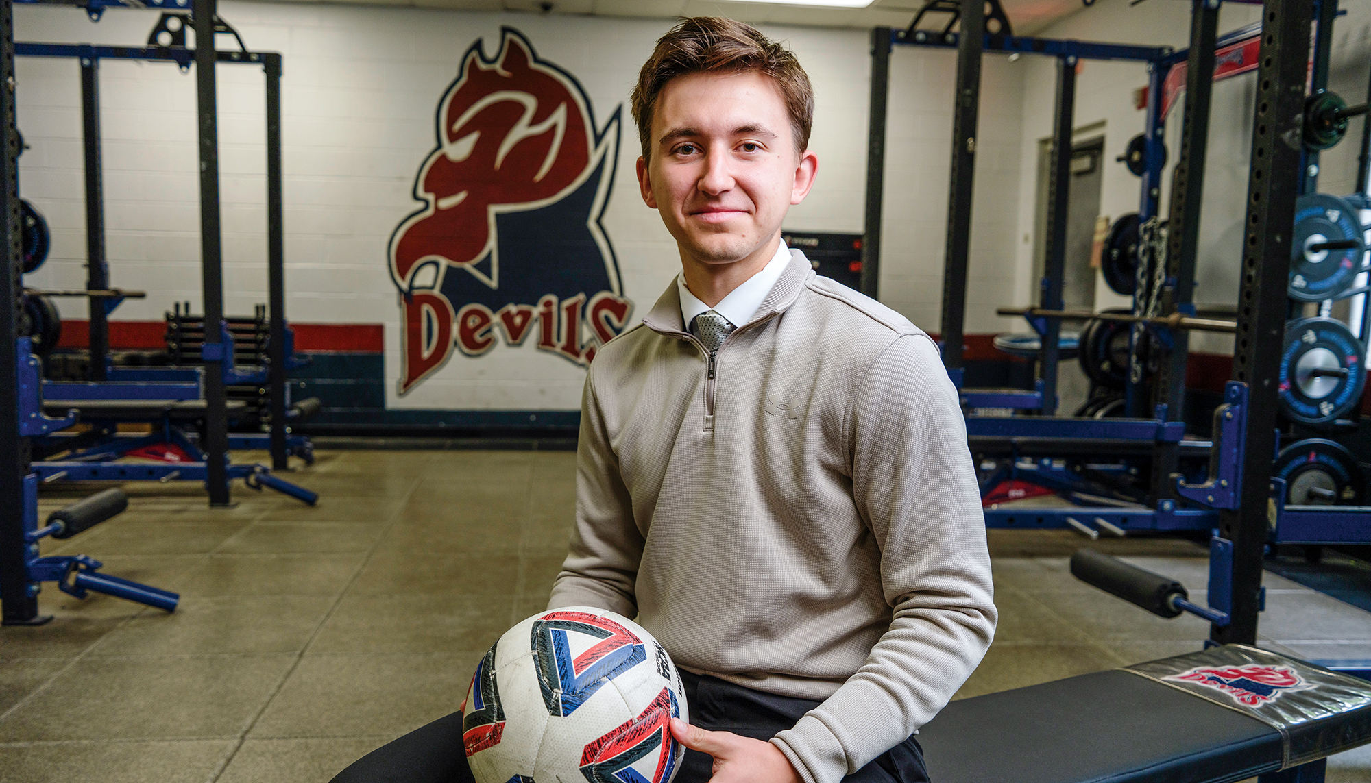 A young man sits in a gym weight room, holding a soccer ball.
