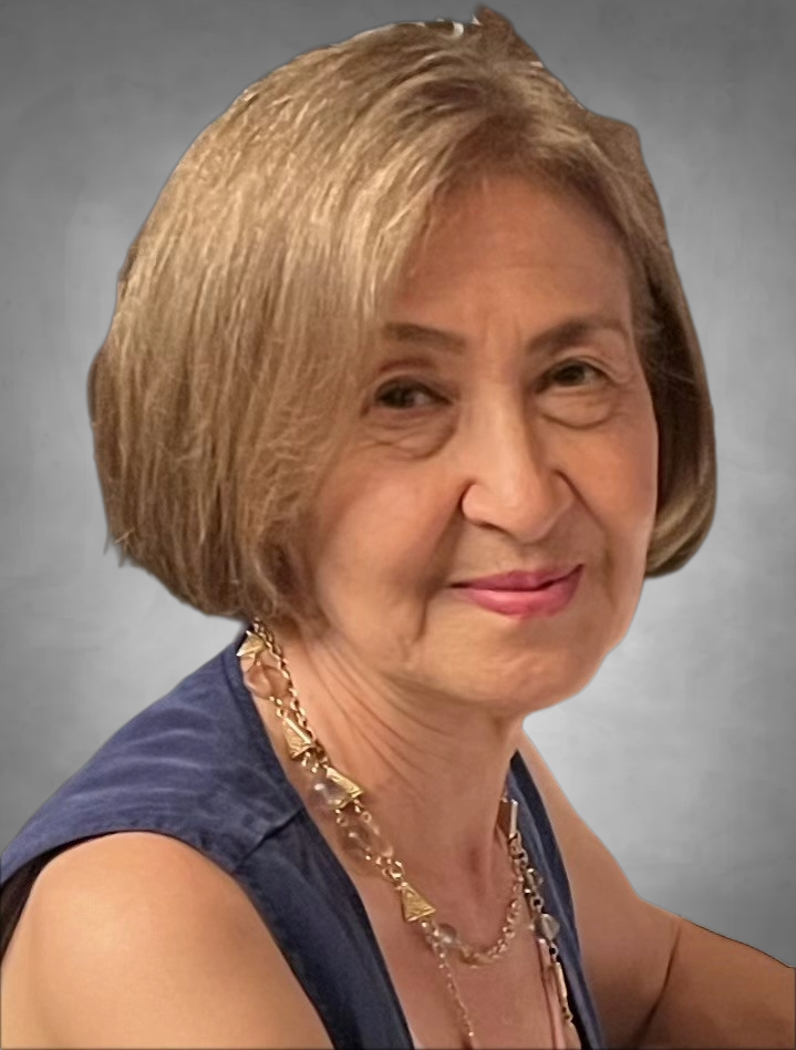 A professional headshot of a woman with short blonde hair styled in a bob. She is wearing a navy blue sleeveless top and layered gold necklaces. She has a warm, gentle smile and is photographed against a neutral gray background.