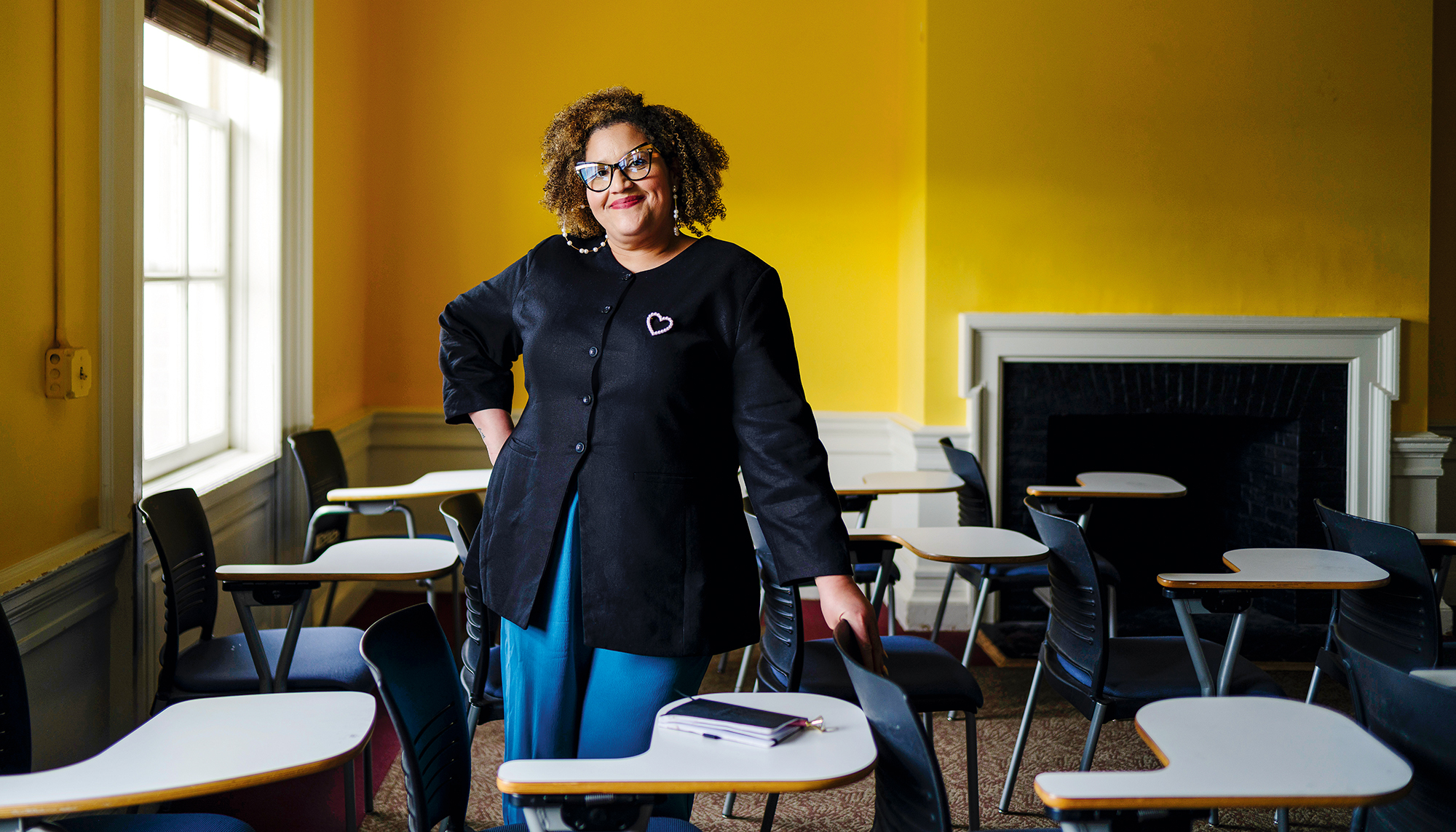A woman wearing glasses, smiles and puts her hands on her hip in a classroom.