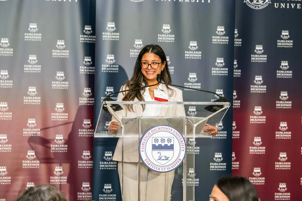 A woman wearing a tan suit stands at a podium giving a speech surrounded by FDU banners.