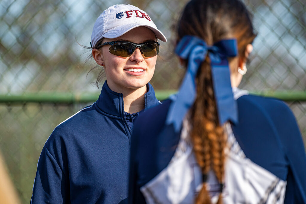 A woman wearing a baseball hat and sunglasses looks on at a student-athlete.