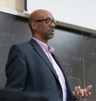 a professor stands at a chalkboard and speaks to a class out of frame.