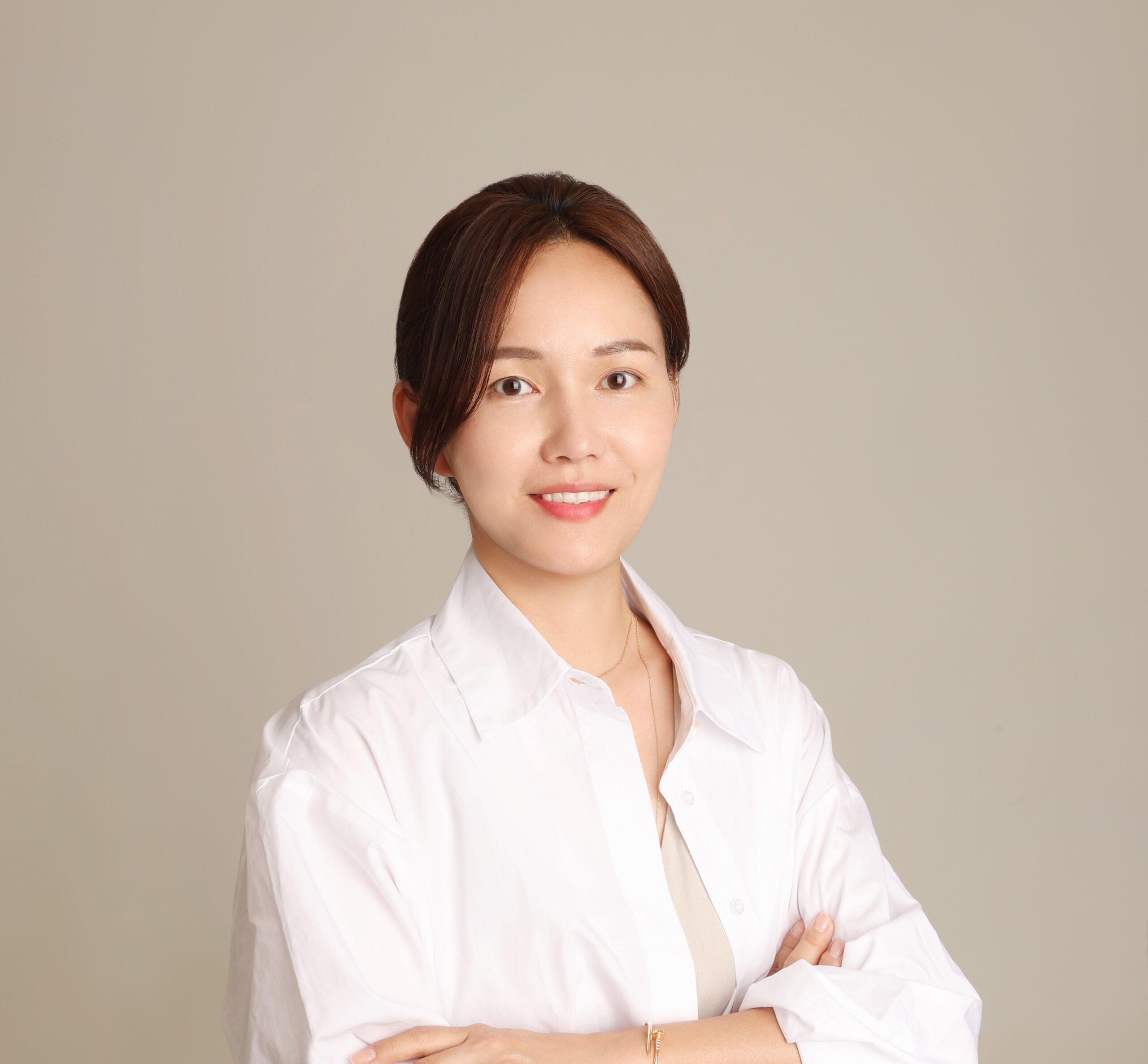 A professional headshot of a woman with dark hair pulled back, wearing a white collared blouse. She has her arms crossed and is smiling warmly at the camera against a neutral beige background.