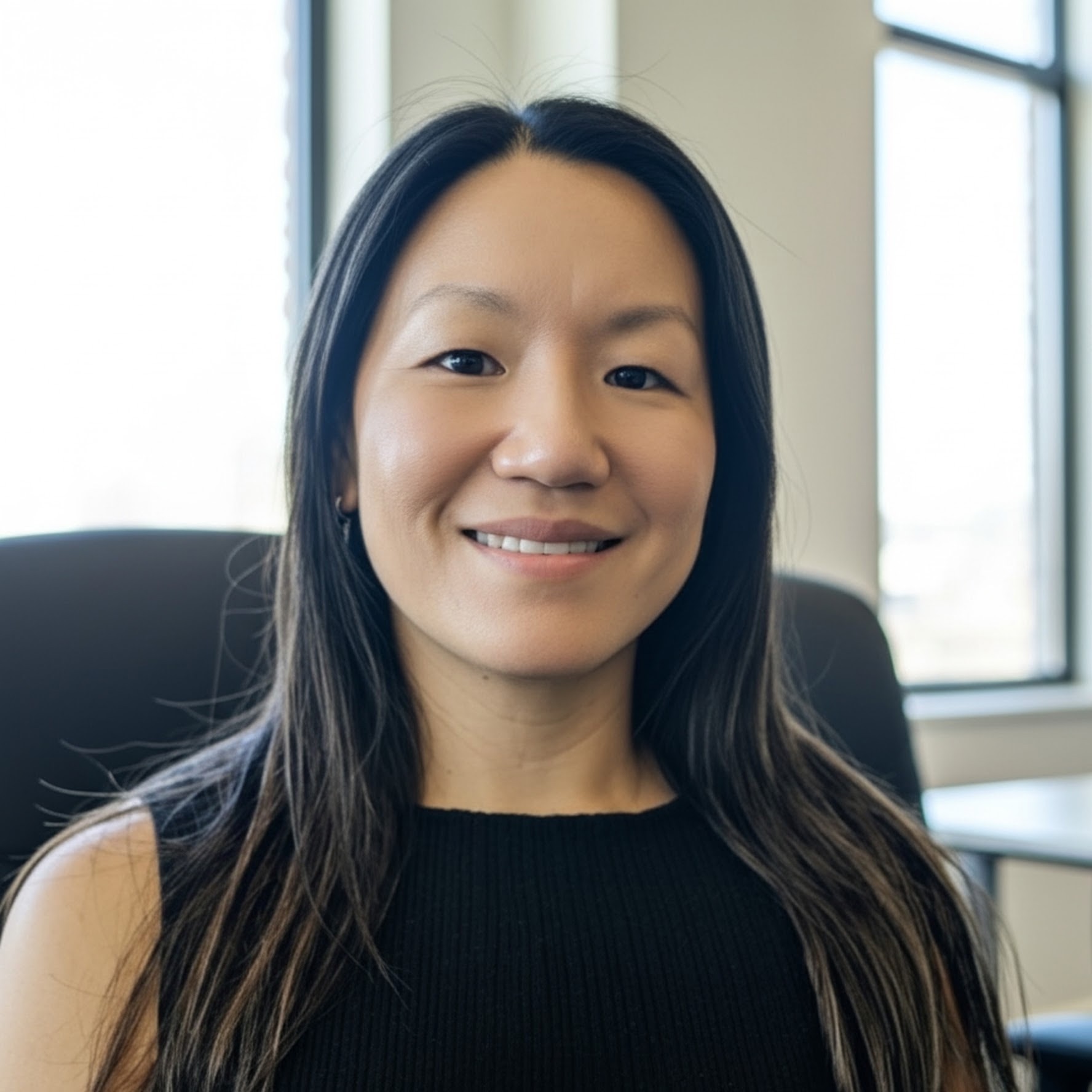 A professional headshot of a woman with long dark hair wearing a black sleeveless top. She is seated in an office chair with natural light from windows in the background, smiling at the camera.