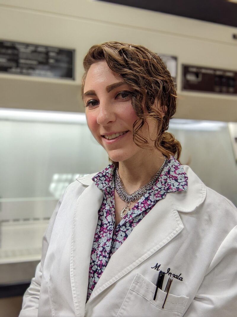 A professional photo of a person with wavy brown hair pulled back, standing in a laboratory setting. They are wearing a white lab coat with an embroidered name and pens in the pocket, over a floral patterned blouse with purple and pink flowers. They are wearing a silver chain necklace and smiling at the camera.